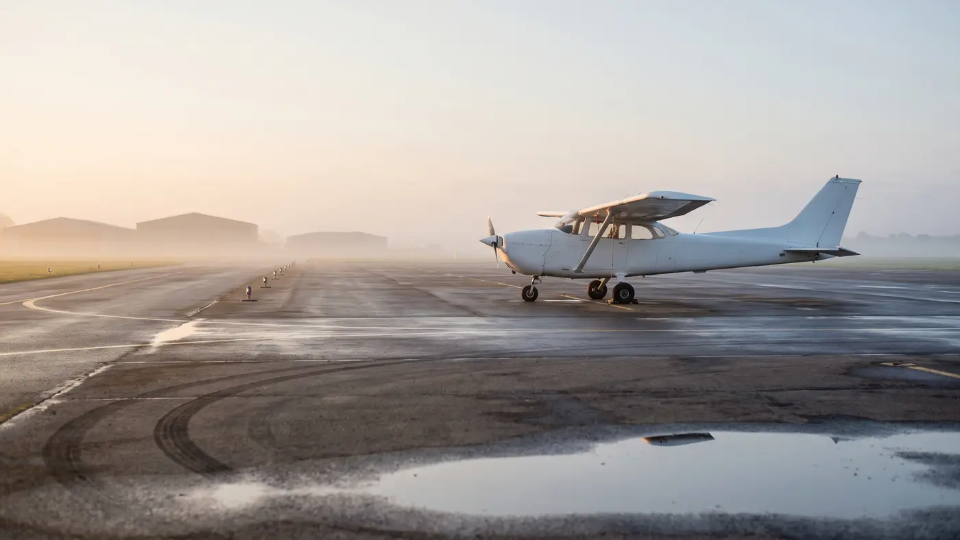 A small unmarked training aircraft parked on a wet airfield apron at dawn, runway lights receding into mist.